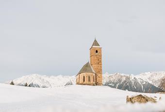 Die St. Kathrein Kirche in der Scharte in Hafling umgeben von frischem pulverschnee. Im Hintergrund die weißen Bergspitzen im Winter. Bisschen vor der Kirche der markante Stein.