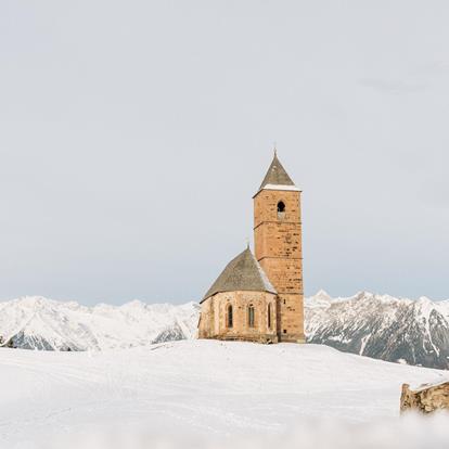 The St. Kathrein church in the Scharte in Hafling surrounded by fresh powder snow. In the background the white mountain peaks in winter. The striking stone in front of the church.